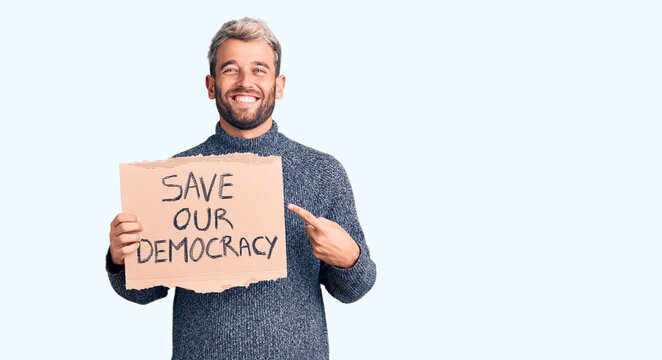 Young blond man holding save our democracy cardboard banner smiling happy pointing with hand and finger