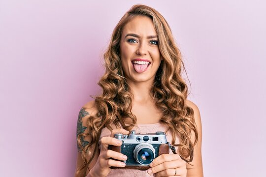 Young blonde girl holding vintage camera sticking tongue out happy with funny expression.