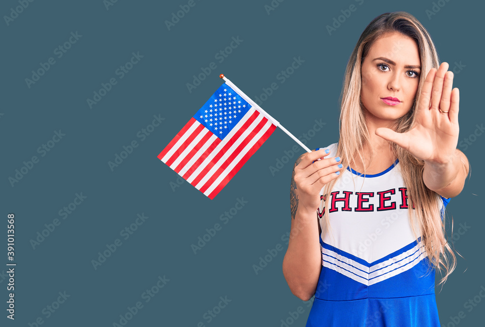 Wall mural Young beautiful blonde woman wearing cheerleader uniform holding united states flag with open hand doing stop sign with serious and confident expression, defense gesture