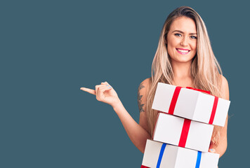 Young beautiful blonde woman holding birthday gifts smiling happy pointing with hand and finger to the side