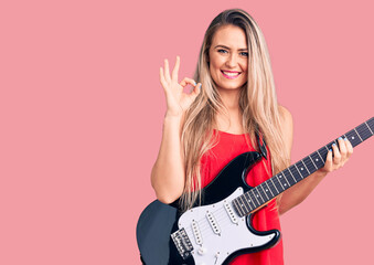 Fototapeta premium Young beautiful blonde woman playing electric guitar doing ok sign with fingers, smiling friendly gesturing excellent symbol