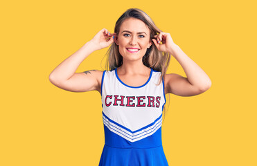 Young beautiful blonde woman wearing cheerleader uniform smiling pulling ears with fingers, funny...