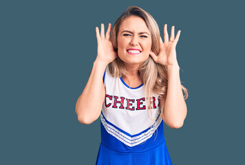 Young beautiful blonde woman wearing cheerleader uniform trying to hear both hands on ear gesture, curious for gossip. hearing problem, deaf