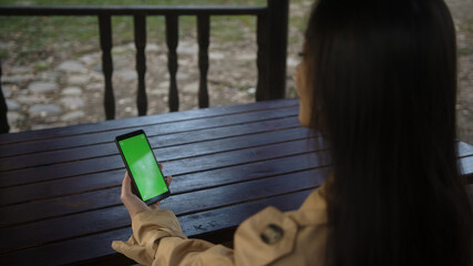 Young woman at nature sitting on a bench using with Green Mock-up Screen Smartphone in Horizontal Landscape Mode. Girl Using Mobile Phone, Browsing Internet, Watching Content