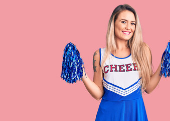 Young beautiful blonde woman wearing cheerleader uniform holding pompom looking positive and happy...