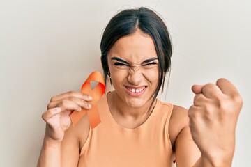 Young brunette woman holding awareness orange ribbon annoyed and frustrated shouting with anger,...