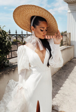 Beautiful Woman With Dark Hair In Luxurious Wedding Dress And Big Straw Hat Posing At The Balcony