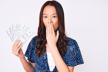 Young beautiful chinese girl holding dollars covering mouth with hand, shocked and afraid for...