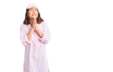 Young beautiful chinese girl wearing sleep mask and pajama begging and praying with hands together...