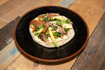 A delicious plate of a naan flatbread with Hummus, Mushrooms, Serrano Ham and Avocado on a wooden kitchen work top