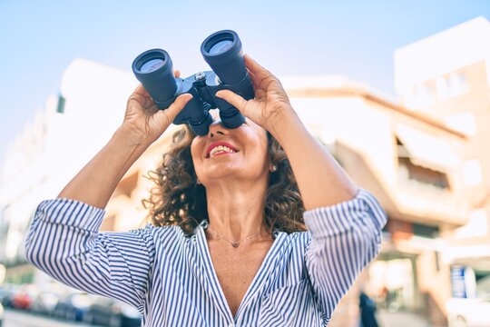 Middle age hispanic woman smiling happy looking for new opportunity using binoculars at the city.