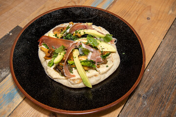 A delicious plate of a naan flatbread with Hummus, Mushrooms, Serrano Ham and Avocado on a wooden kitchen work top
