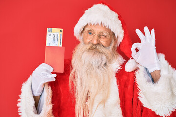 Old senior man with grey hair and long beard wearing santa claus costume holding boarding pass doing ok sign with fingers, smiling friendly gesturing excellent symbol