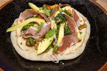 A delicious plate of a naan flatbread with Hummus, Mushrooms, Serrano Ham and Avocado on a wooden kitchen work top