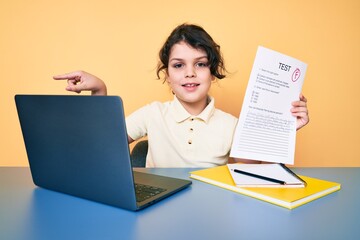 Cute hispanic child showing failed exam sitting on the desk smiling happy pointing with hand and finger to the side