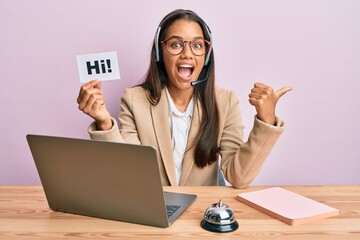 Beautiful hispanic woman wearing operator headset holding hi paper pointing thumb up to the side...