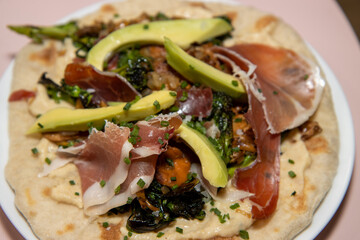 A delicious plate of a naan flatbread with Hummus, Mushrooms, Serrano Ham and Avocado on a wooden kitchen work top