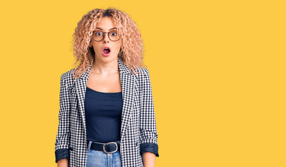 Young blonde woman with curly hair wearing business jacket and glasses afraid and shocked with...