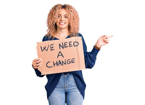 Young blonde woman with curly hair holding we need a change banner smiling happy pointing with hand and finger to the side