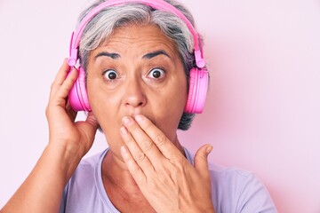 Senior hispanic woman listening to music using headphones covering mouth with hand, shocked and...