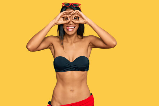Young african american woman wearing bikini doing ok gesture like binoculars sticking tongue out, eyes looking through fingers. crazy expression.