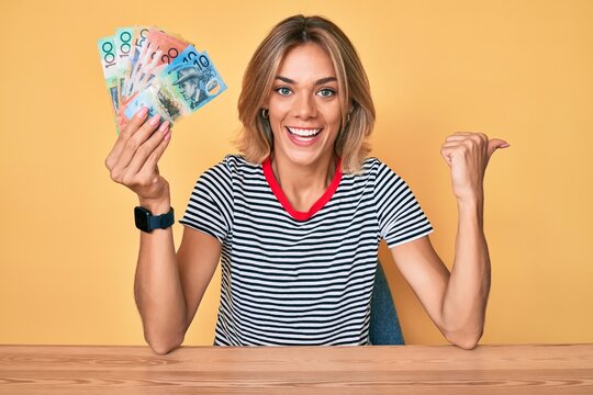 Beautiful caucasian woman holding australian dollars pointing thumb up to the side smiling happy with open mouth