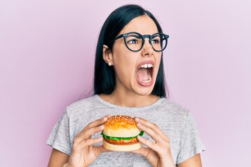 Beautiful young woman eating tasty hamburger angry and mad screaming frustrated and furious, shouting with anger. rage and aggressive concept.