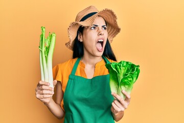 Beautiful young woman wearing gardener apron holding vegetables angry and mad screaming frustrated...
