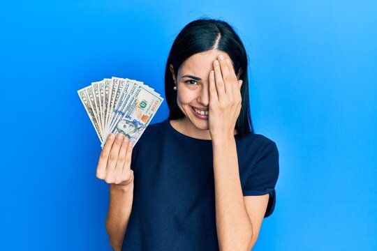 Beautiful young woman holding dollars covering one eye with hand, confident smile on face and surprise emotion.