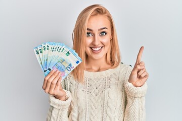 Beautiful caucasian blonde woman holding 20 euro banknotes smiling with an idea or question pointing finger with happy face, number one