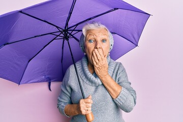 Senior grey-haired woman holding purple umbrella covering mouth with hand, shocked and afraid for...