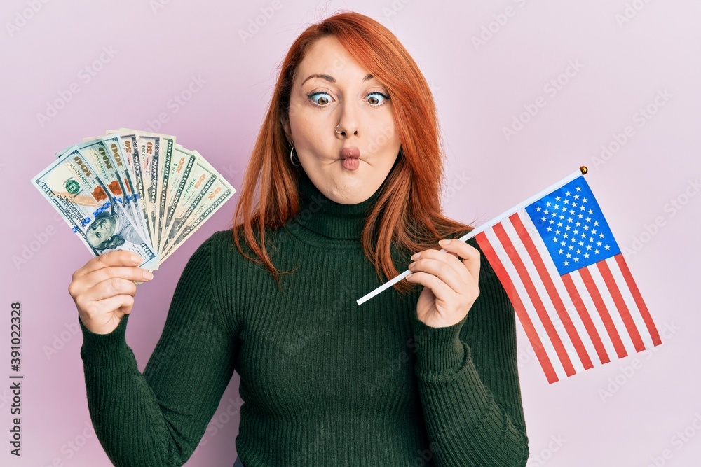 Poster beautiful redhead woman holding dollars banknote and usa flag making fish face with mouth and squint