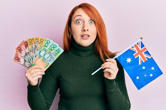 Beautiful redhead woman holding australian dollars and australia flag afraid and shocked with surprise and amazed expression, fear and excited face.
