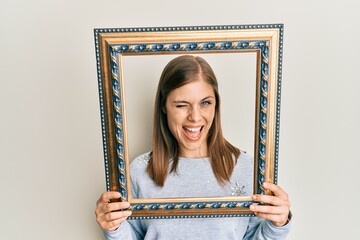 Beautiful caucasian woman holding empty frame winking looking at the camera with sexy expression,...