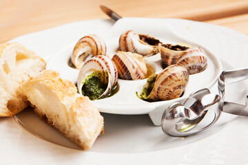 snail dish, served on a white classic plate, with various sauces (pesto, truffle, cheese), pieces of French baguette, fork for a shell, wooden table, from close range, space, horizontal, no one

