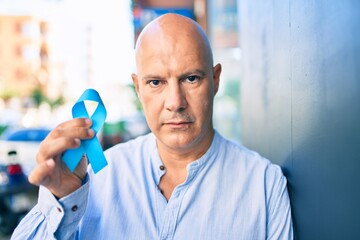 Middle age bald man with serious expression holding blue prostate cancer ribbon leaning on the wall at the city
