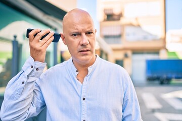 Middle age bald man with serious expression listening audio message using smartphone at the city.