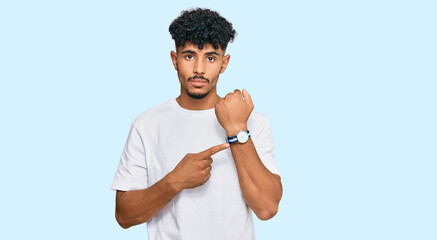 Young arab man wearing casual white t shirt in hurry pointing to watch time, impatience, looking at...