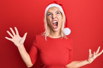 Young blonde girl wearing christmas hat crazy and mad shouting and yelling with aggressive expression and arms raised. frustration concept.