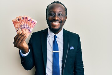 Handsome young black man holding south african rand banknotes looking positive and happy standing and smiling with a confident smile showing teeth