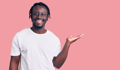 Young african american man with braids wearing casual white tshirt smiling cheerful presenting and pointing with palm of hand looking at the camera.