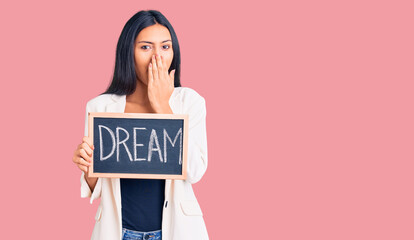 Young beautiful latin girl holding blackboard with dream word covering mouth with hand, shocked and...