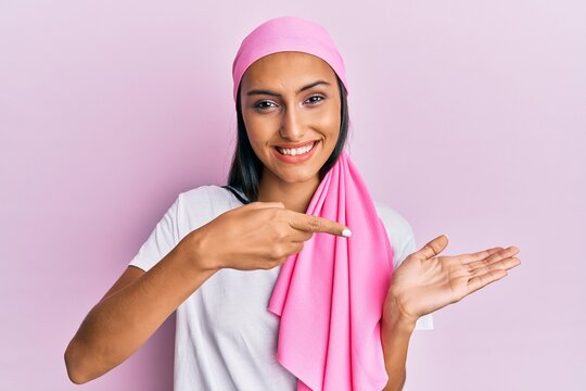 Young brunette woman wearing breast cancer support pink scarf amazed and smiling to the camera while presenting with hand and pointing with finger.