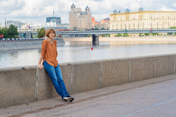 Fototapeta na wymiar An adult redhead woman in casual clothes stands on the embankment, leaning against a stone fence with a strained gaze. River embankment on a warm day.