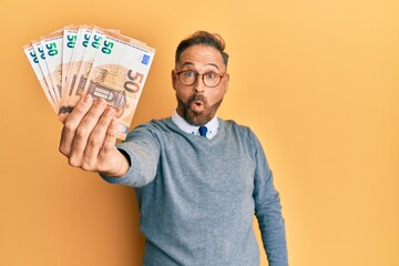 Handsome middle age man holding bunch of euros scared and amazed with open mouth for surprise,...