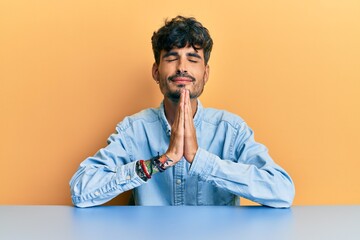 Young hispanic man wearing casual clothes sitting on the table begging and praying with hands...