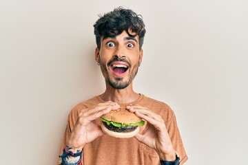 Young hispanic man eating a tasty classic burger celebrating crazy and amazed for success with open...