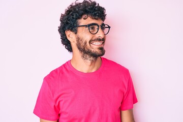 Handsome young man with curly hair and bear wearing casual clothes and glasses looking away to side with smile on face, natural expression. laughing confident.