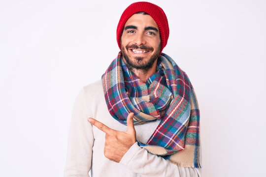 Handsome young man with curly hair and bear wearing a winter sweater, scarf and wool hat smiling cheerful pointing with hand and finger up to the side