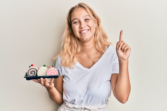 Young blonde girl holding cake sweets smiling with an idea or question pointing finger with happy face, number one